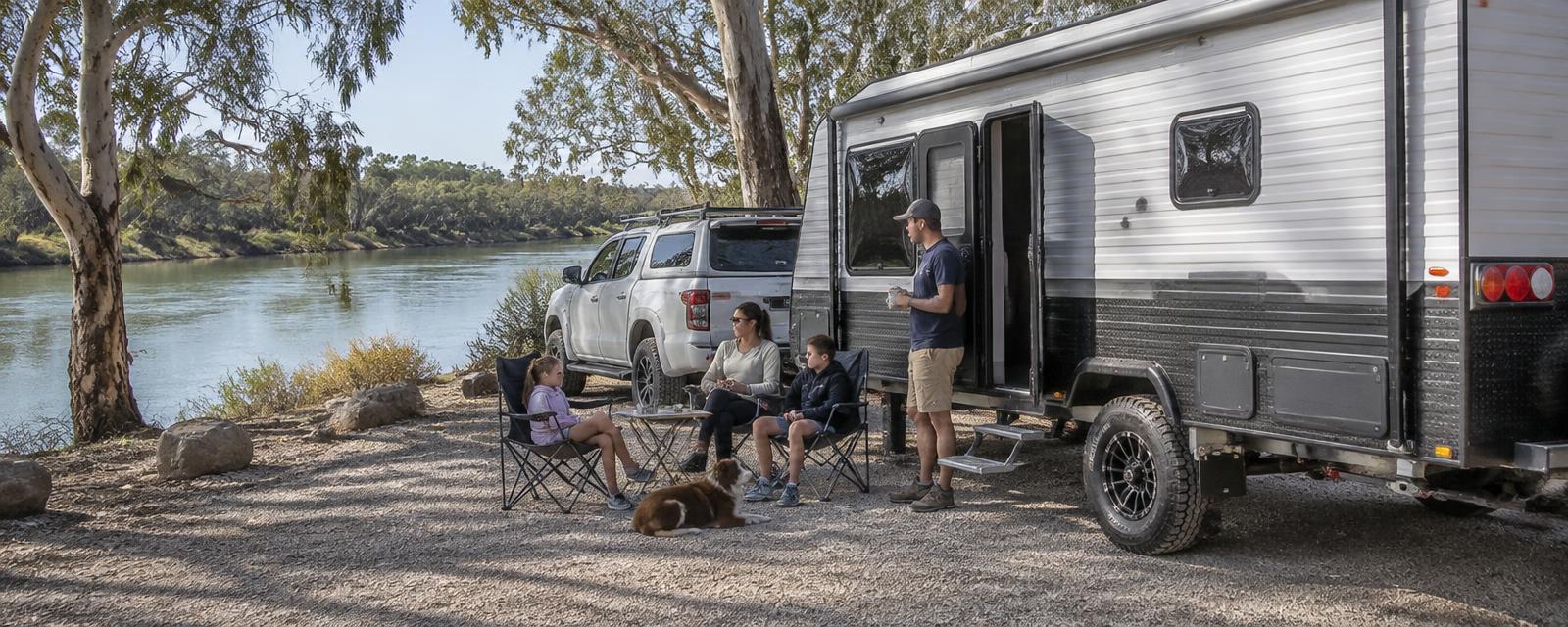Family and their dog sitting on a riverbank beside a caravan and pickup truck at a gravel lookout